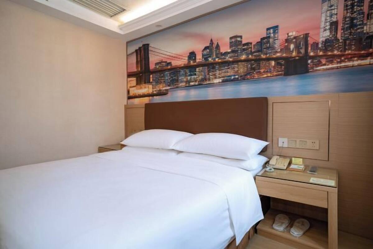 A hotel room with a bed, a nightstand with a phone, and a wall-mounted shelf with slippers.