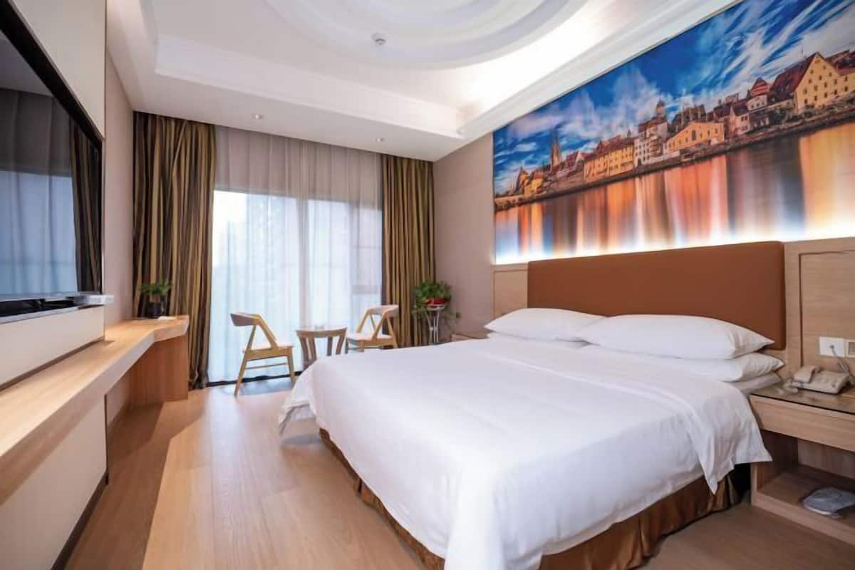 A hotel room with a large bed, a desk with a flat-screen TV, a chair, a small table, and a painting of a cityscape on the wall.