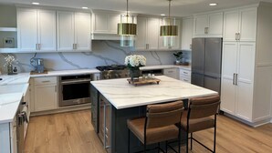Una cucina moderna con mobili bianchi, un'isola in marmo, elettrodomestici in acciaio inossidabile e illuminazione a sospensione.