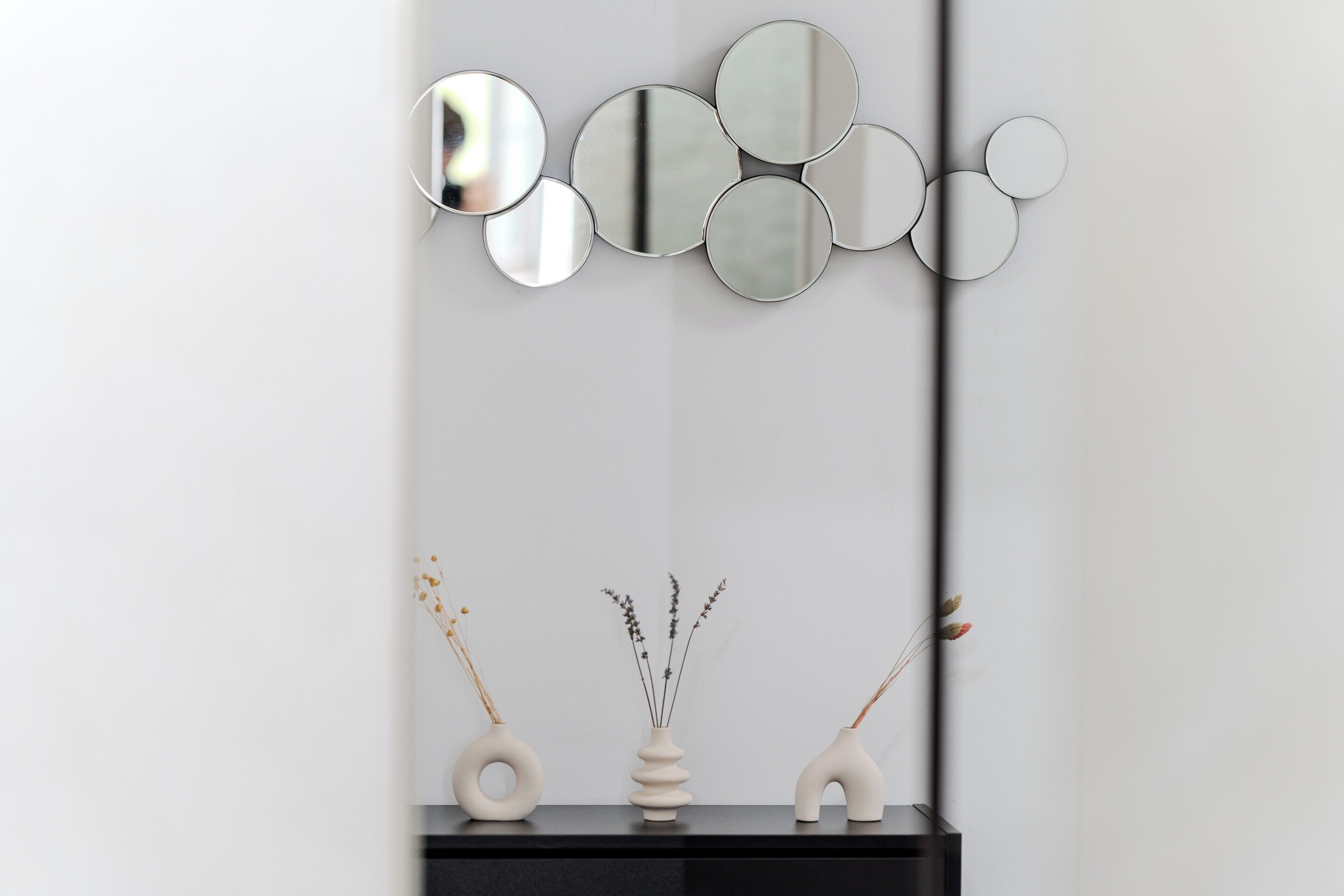 A wall with a series of circular mirrors and three vases with dried plants on a black console.
