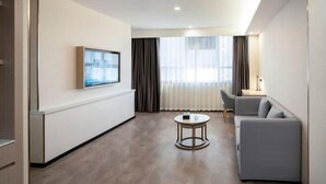 Quarto de hotel moderno com televisão de tela plana na parede, sofá cinza, mesa de centro redonda e uma janela grande com cortinas leves.