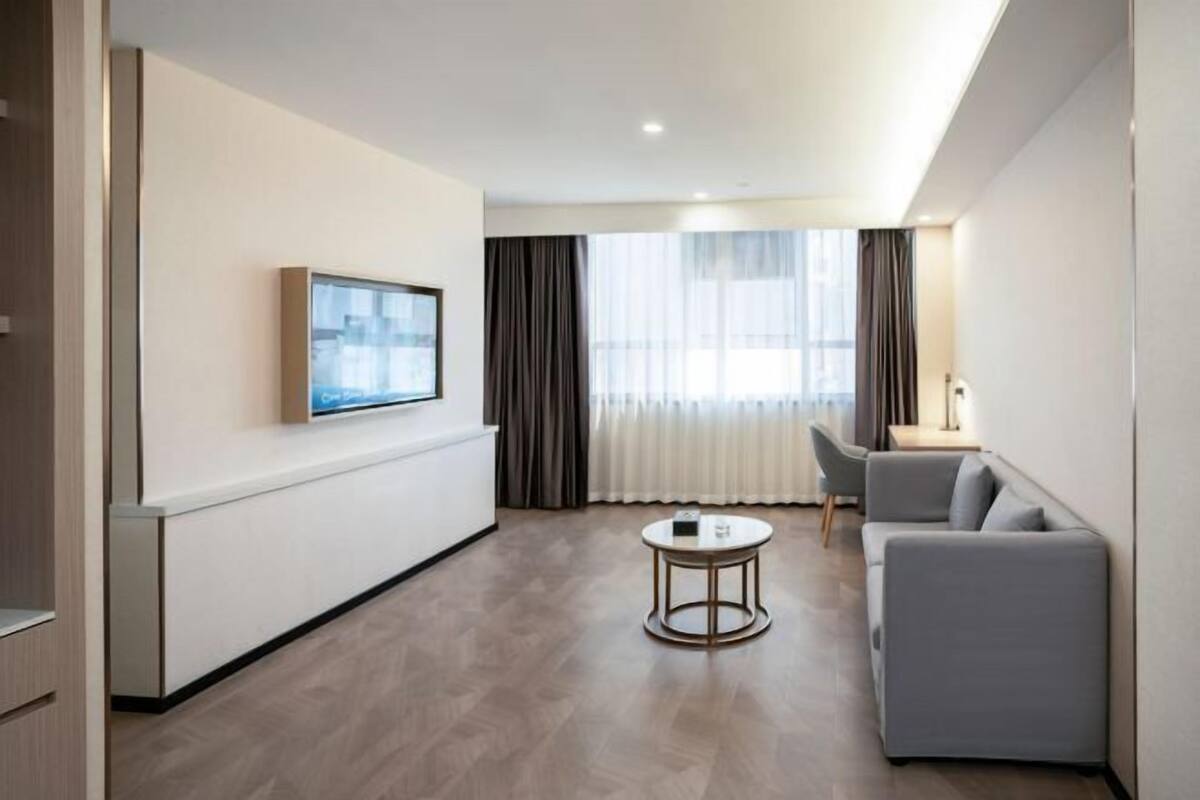 A modern hotel room with a flat-screen TV mounted on the wall, a grey sofa, a small round coffee table, and a large window with sheer curtains.