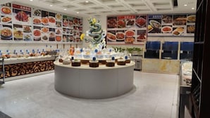 Interior de restaurante com um balcão central redondo exibindo diversos alimentos, rodeado por paredes com quadros de menu.