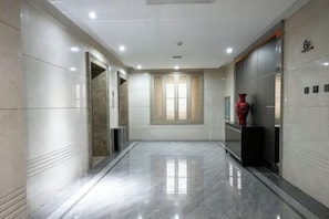 A hallway with a polished floor, a window with blinds, and a red vase on a stand.