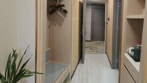 Um corredor estreito com uma planta à esquerda, uma porta no final e uma prateleira com uma chaleira à direita.