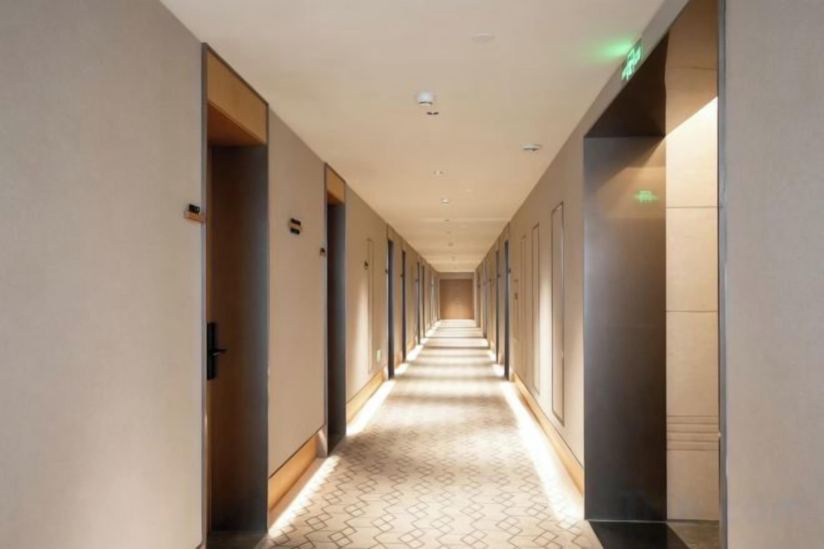 A hotel corridor with doors on both sides, a patterned floor, and a ceiling with recessed lighting.