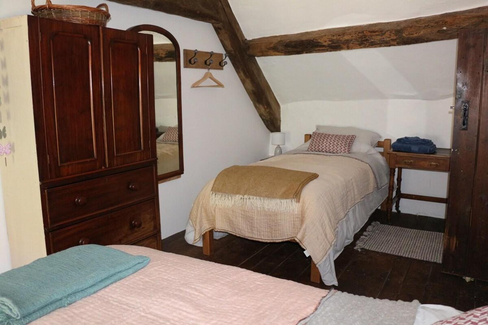 A bedroom with a wooden wardrobe, a bed with a beige blanket, a bedside table with a lamp, and a wooden ceiling beam.