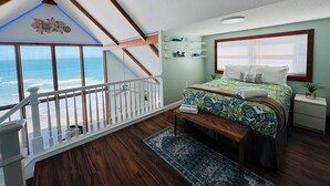 Un dormitorio con vista al océano, vigas de madera, una cama con colcha tropical, una mesita de noche y un pequeño banco.