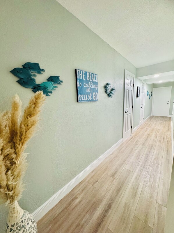 A hallway with a light green wall, wooden flooring, and decorative wall art including a sign that reads "The Beach is calling and I must go."