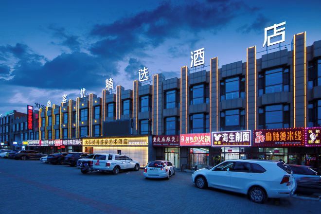 A row of modern buildings with illuminated signage, likely commercial establishments, and a parking lot with several cars.
