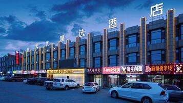 A row of modern buildings with illuminated signage, likely commercial establishments, and a parking lot with several cars.
