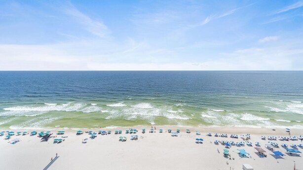 A beach with rows of umbrellas and beach chairs, a sandy shore, and the ocean in the background. - Beach Colony West 13C (Pensacola)