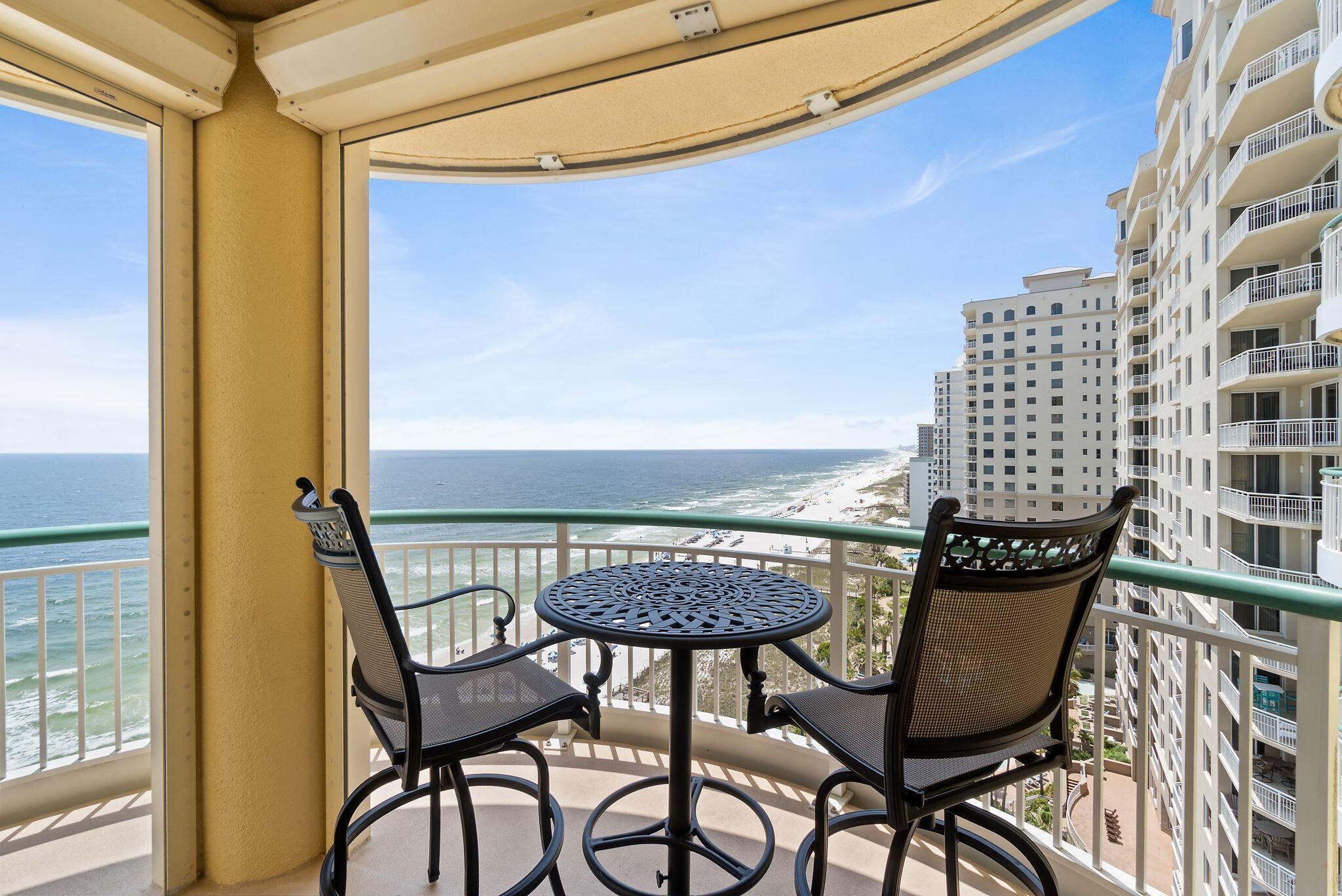 A balcony with a round table and two chairs, overlooking a beach and high-rise buildings.