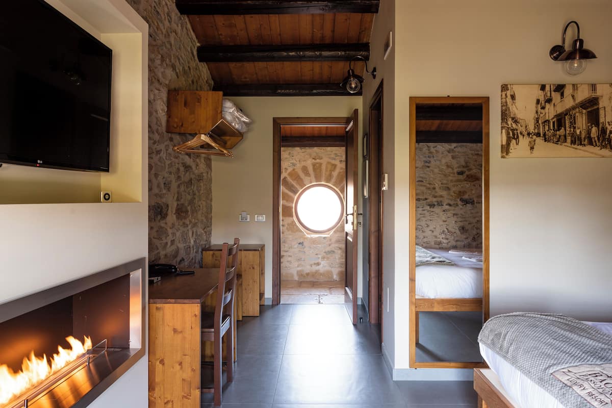 A narrow hallway with a stone wall, a wooden ceiling, a TV mounted on the wall, a fireplace, a bed with a wooden headboard, and a round window.