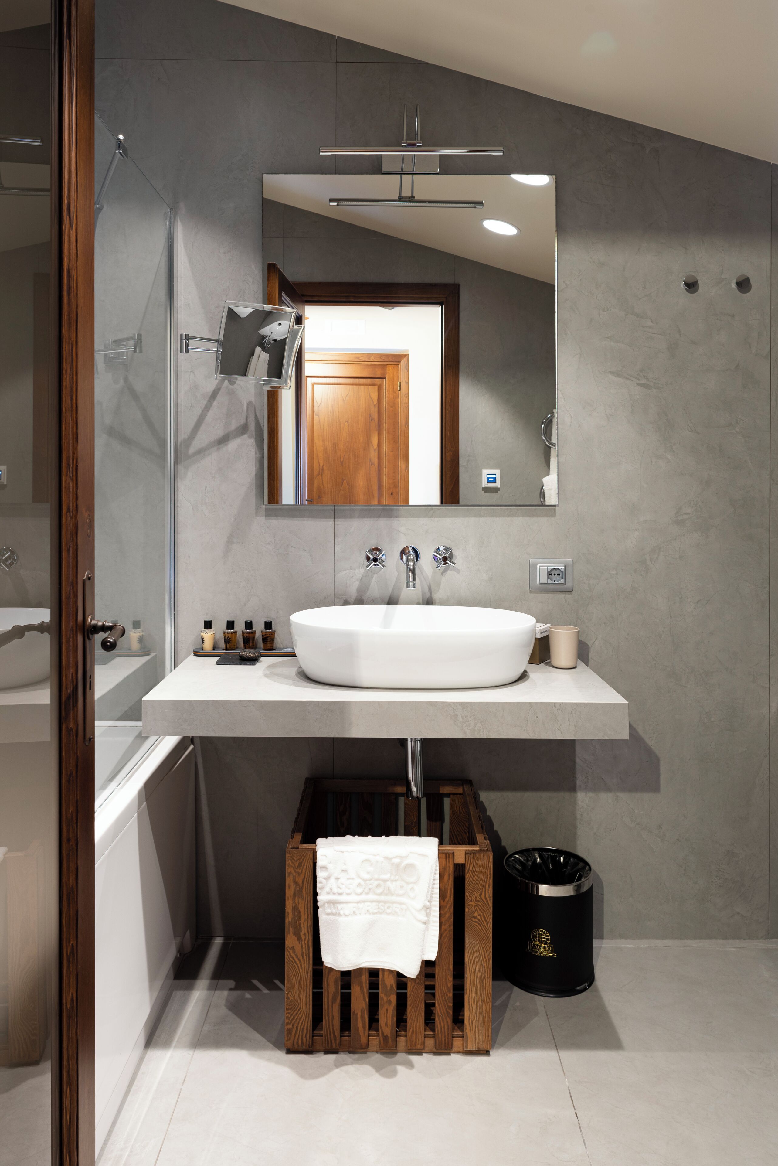 A modern bathroom with a large white sink, a mirror above it, and a wooden crate with a towel.