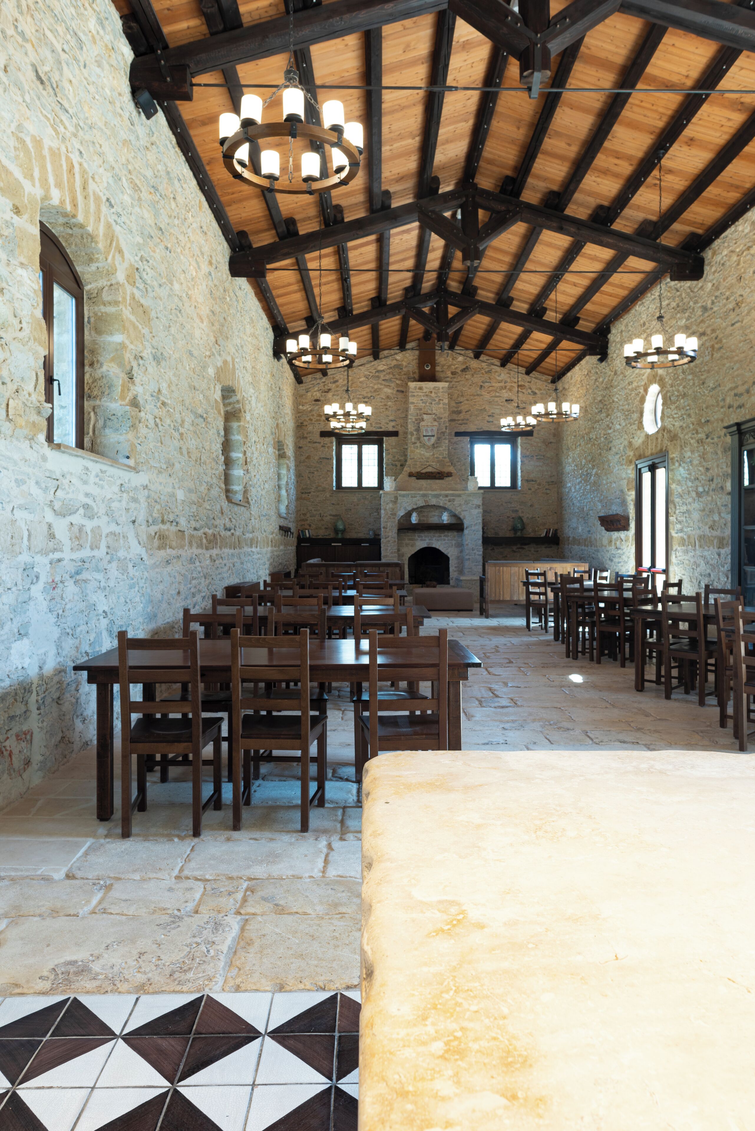A rustic dining hall with stone walls, wooden tables and chairs, and a fireplace.