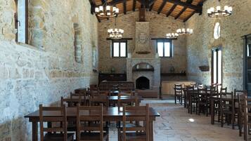A rustic dining hall with stone walls, wooden tables and chairs, and a fireplace.