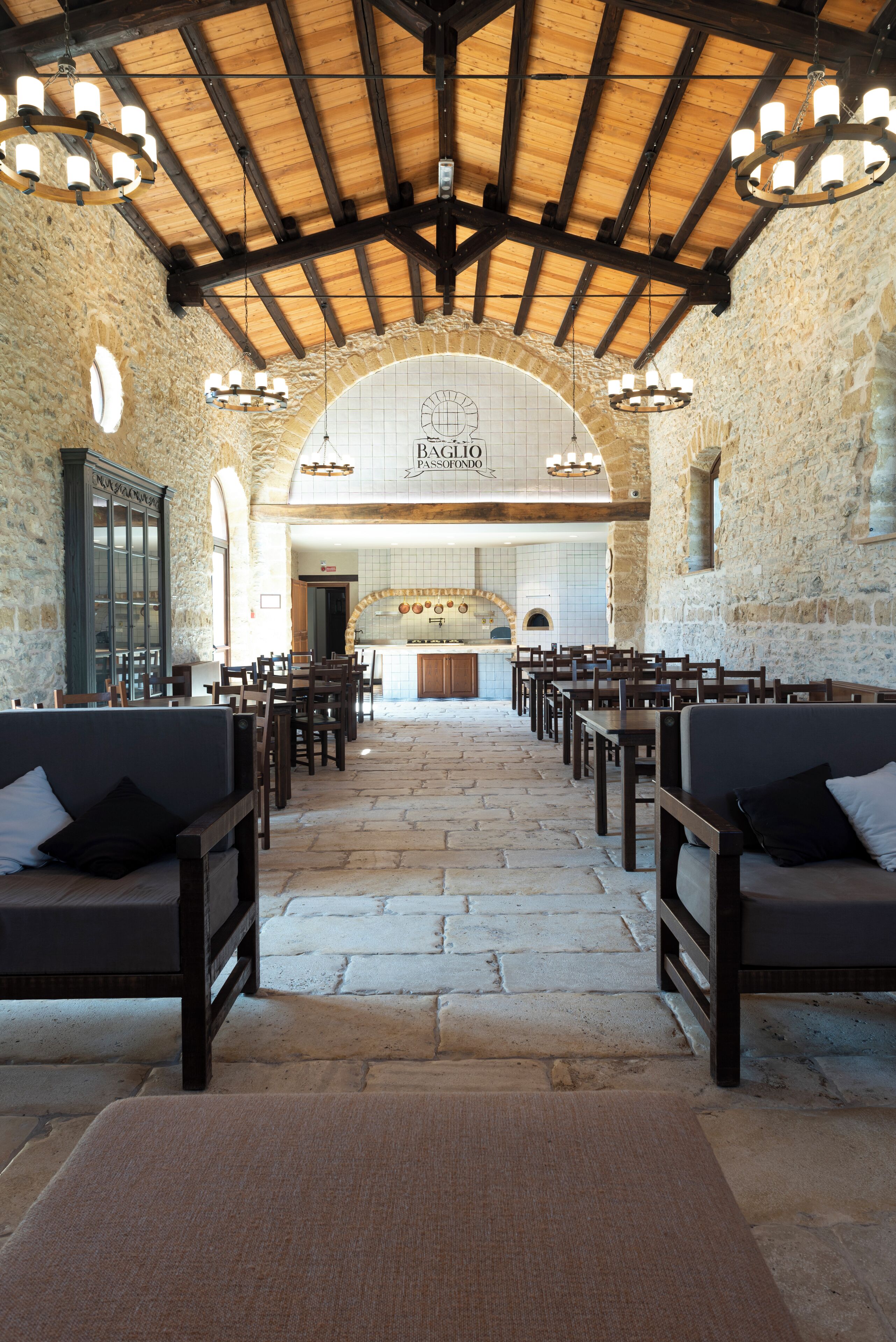 A spacious hall with stone walls and wooden beams, featuring a central kitchen area and multiple seating arrangements.