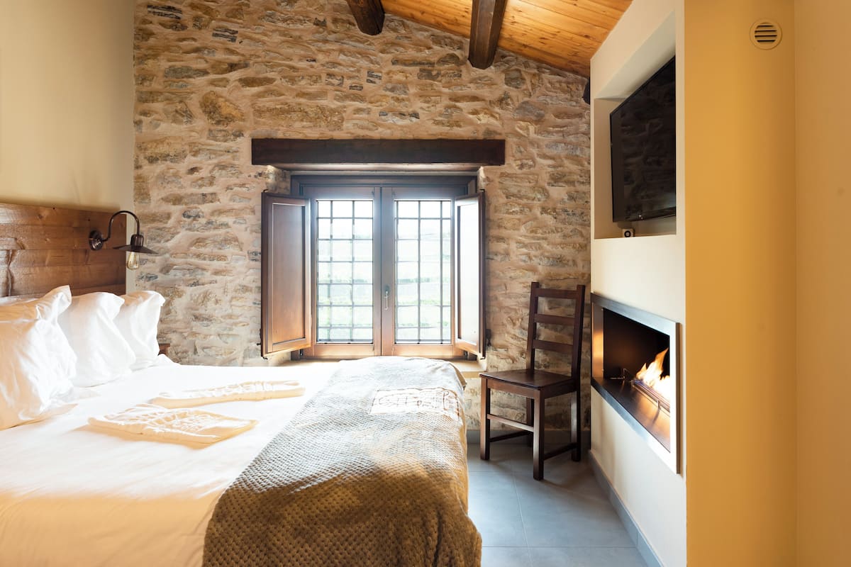 A bedroom with a stone wall, a large bed, a fireplace, and a window with shutters.