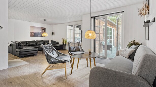 Un salon moderne avec un canapé gris, un divan en cuir noir et un fauteuil en osier. On y trouve une table ronde avec une lampe, ainsi qu’une grande fenêtre donnant sur l’extérieur.