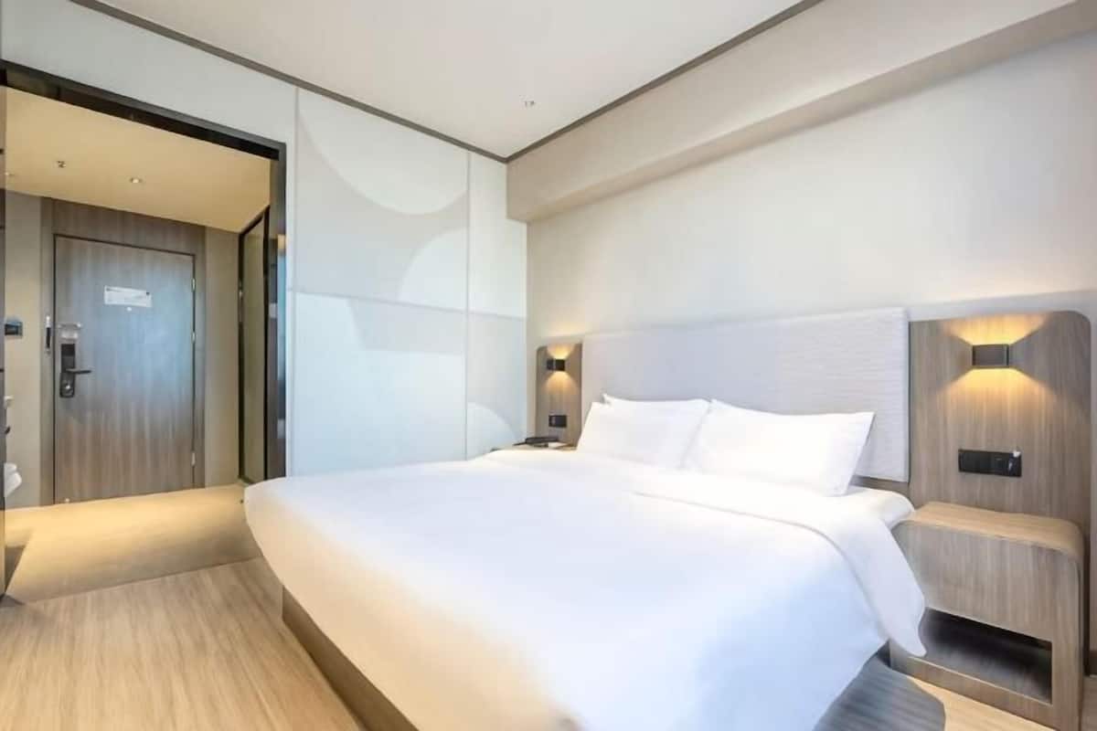 A modern hotel room with a large bed, a wooden door, and a nightstand with a lamp.