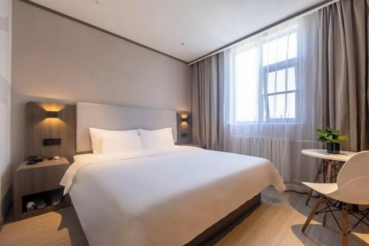 A hotel room with a large bed, bedside tables, a small round table with a chair, and a window with curtains.