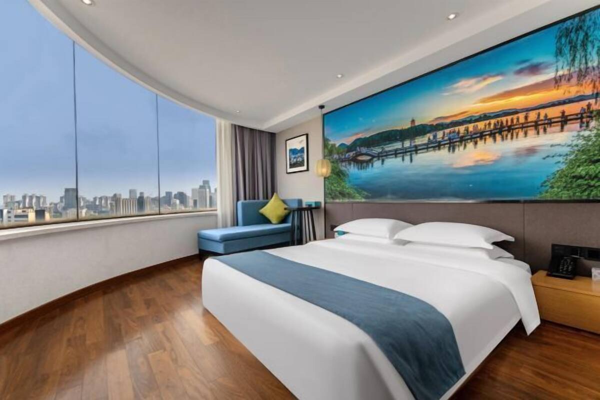 A modern hotel room with a large bed, a city view, a painting of a sunset, and a blue sofa.