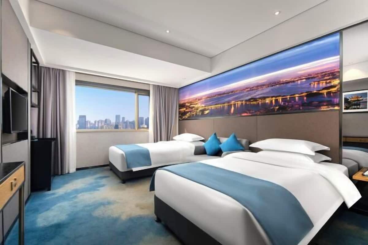 A hotel room with two beds, a large window with a city view, a flat-screen TV, and a painting of a cityscape.