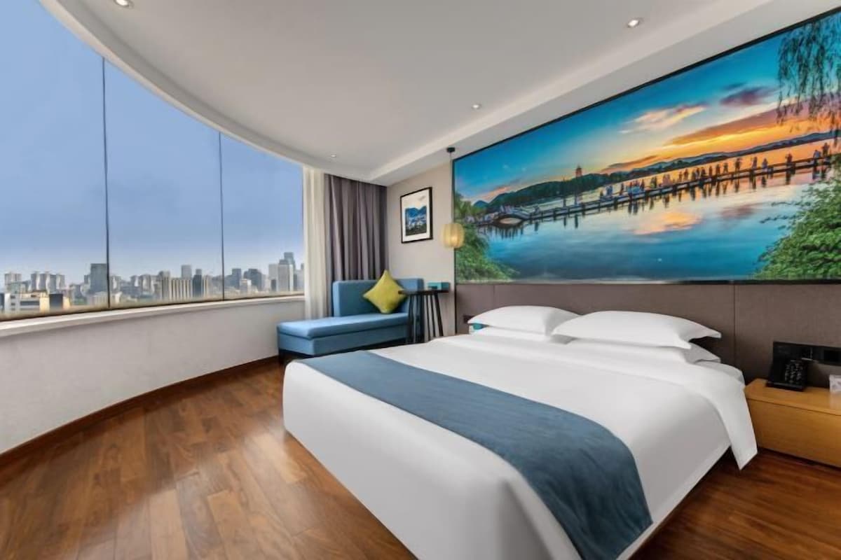 A modern hotel room with a large bed, a city view, a painting on the wall, and a blue sofa.