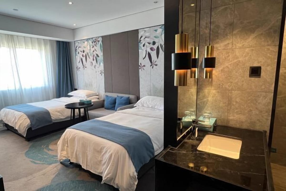 A hotel room with two beds, a sitting area, a large mirror, and a modern bathroom with a marble sink.