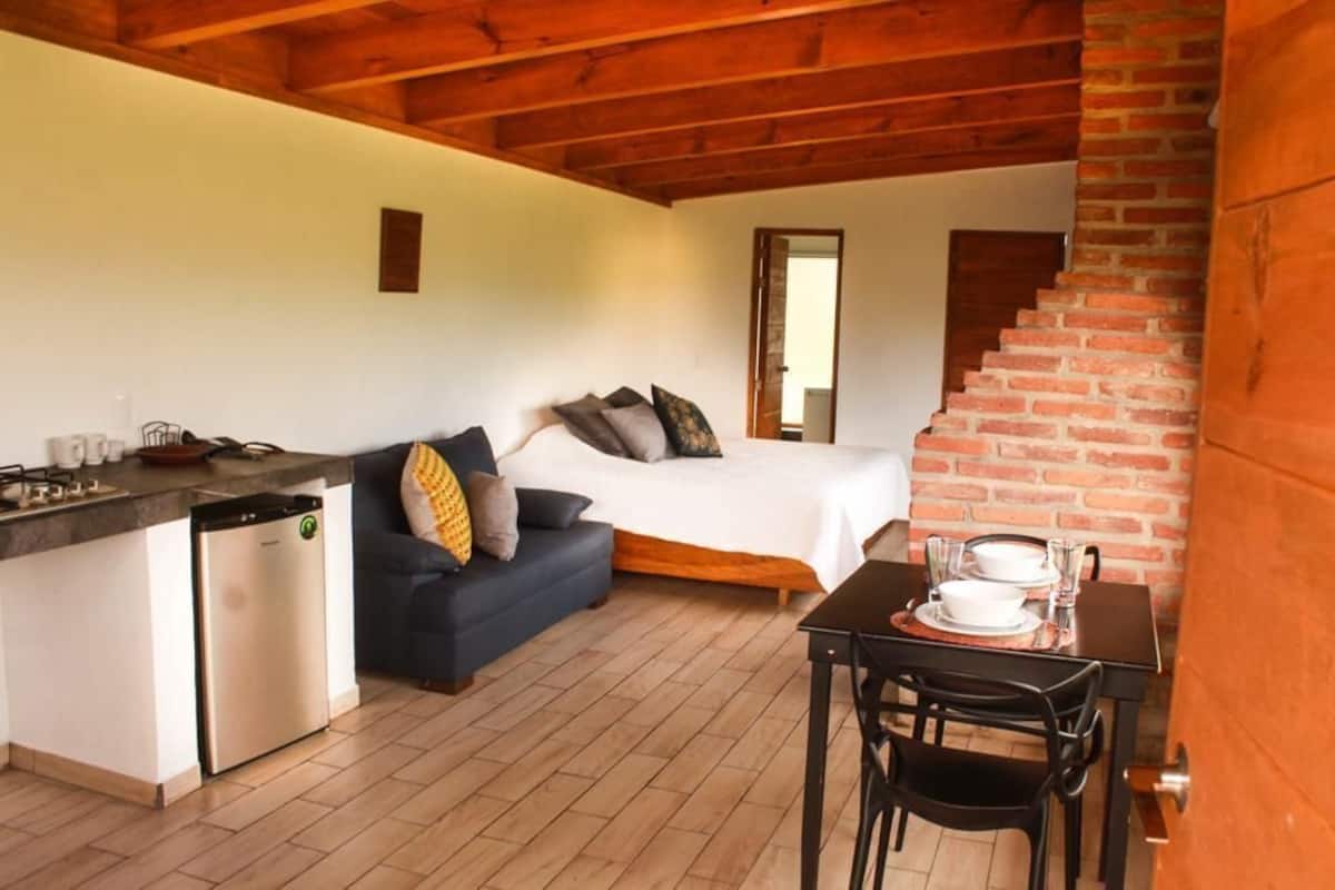 A compact room with a bed, a sofa, a dining table with chairs, a kitchen area with a dishwasher, and a brick staircase.