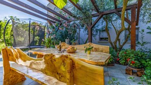 A wooden bench and table set in a covered outdoor area with a pergola structure, surrounded by greenery and potted plants. - Stunning home in Liznjan with private swimming pool, can be inside or outside (Liznjan)