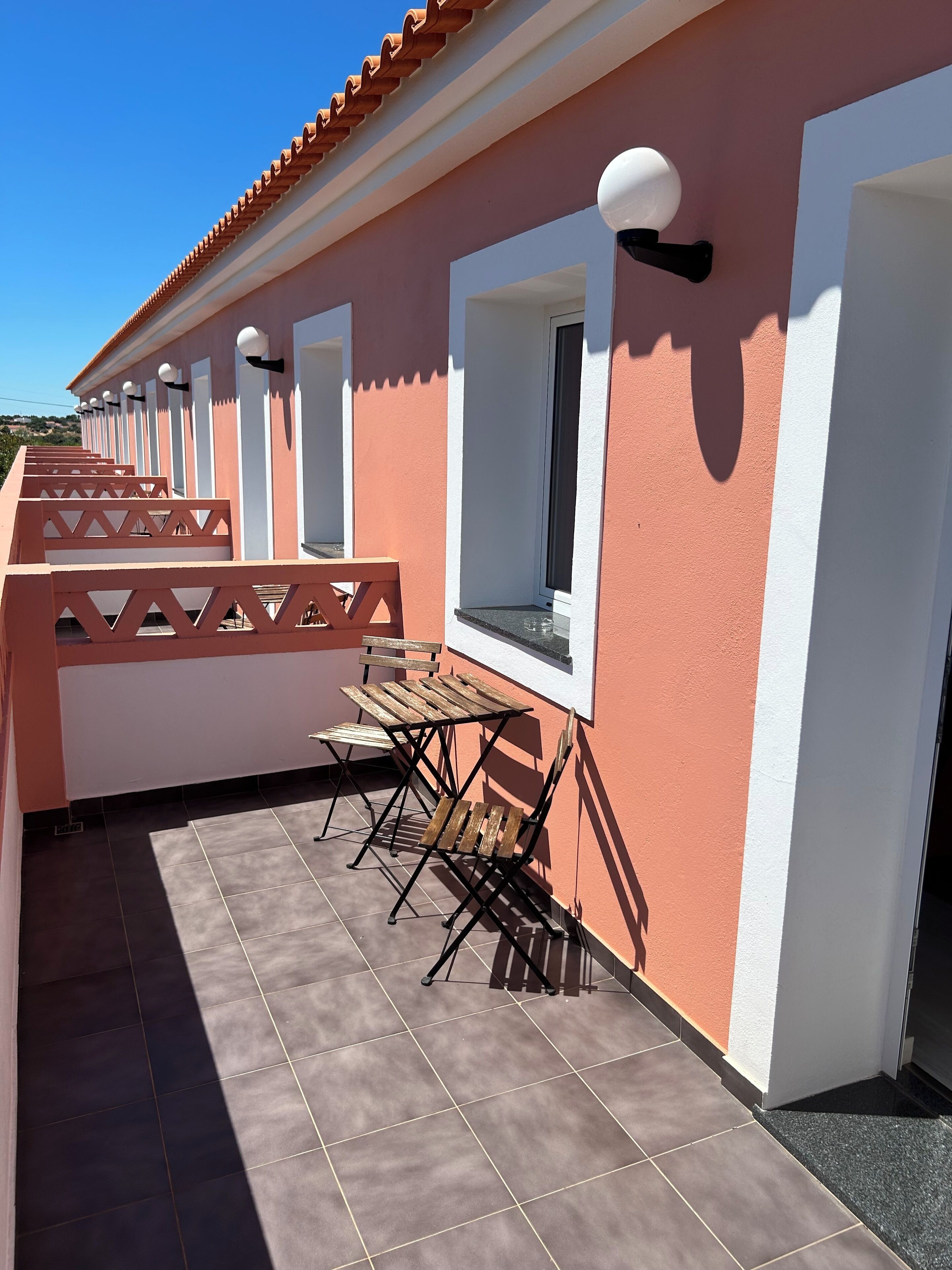 Un espace extérieur abrité, aménagé avec une table et des chaises, doté d’un sol carrelé et bordé d’un bâtiment à la façade rouge et blanche.