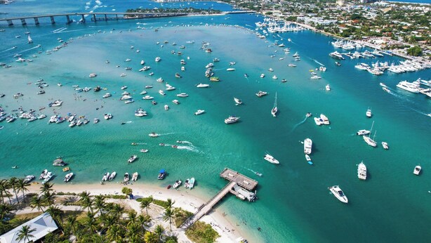An aerial view of a marina with numerous boats docked, a bridge in the background, and a beach area with palm trees.