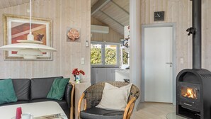 Et hyggeligt rum med sofa, en fletstol, et bord med en kop, en pejs og et vindue med udsigt over grønne omgivelser.
