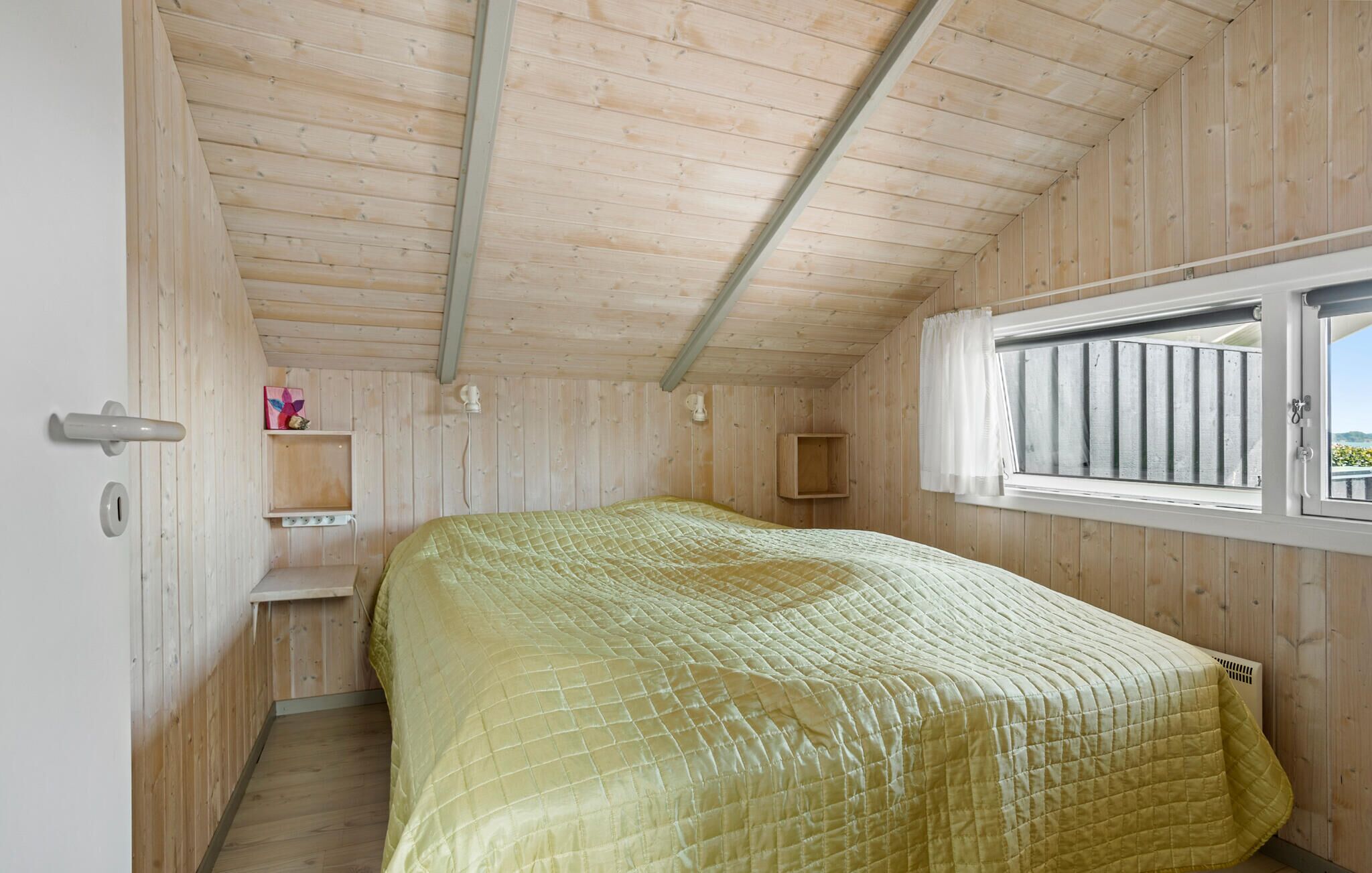 A bedroom with a wooden ceiling, a bed with a quilted bedspread, a window with a view, and a small shelf with items.