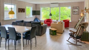 A cozy living room with a dining area, wooden floors, and large windows offering a view of greenery.