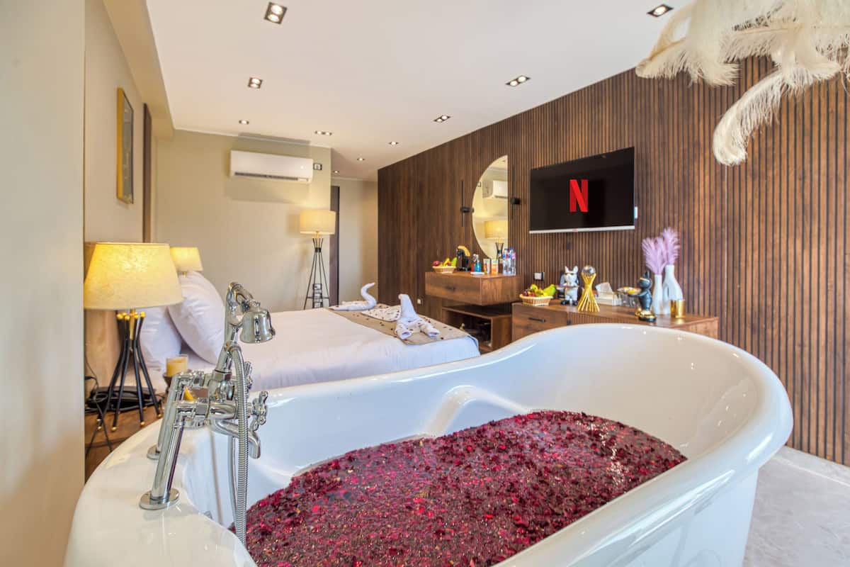 A hotel room with a large bathtub filled with rose petals, a bed, a desk with a TV, and a mirror.