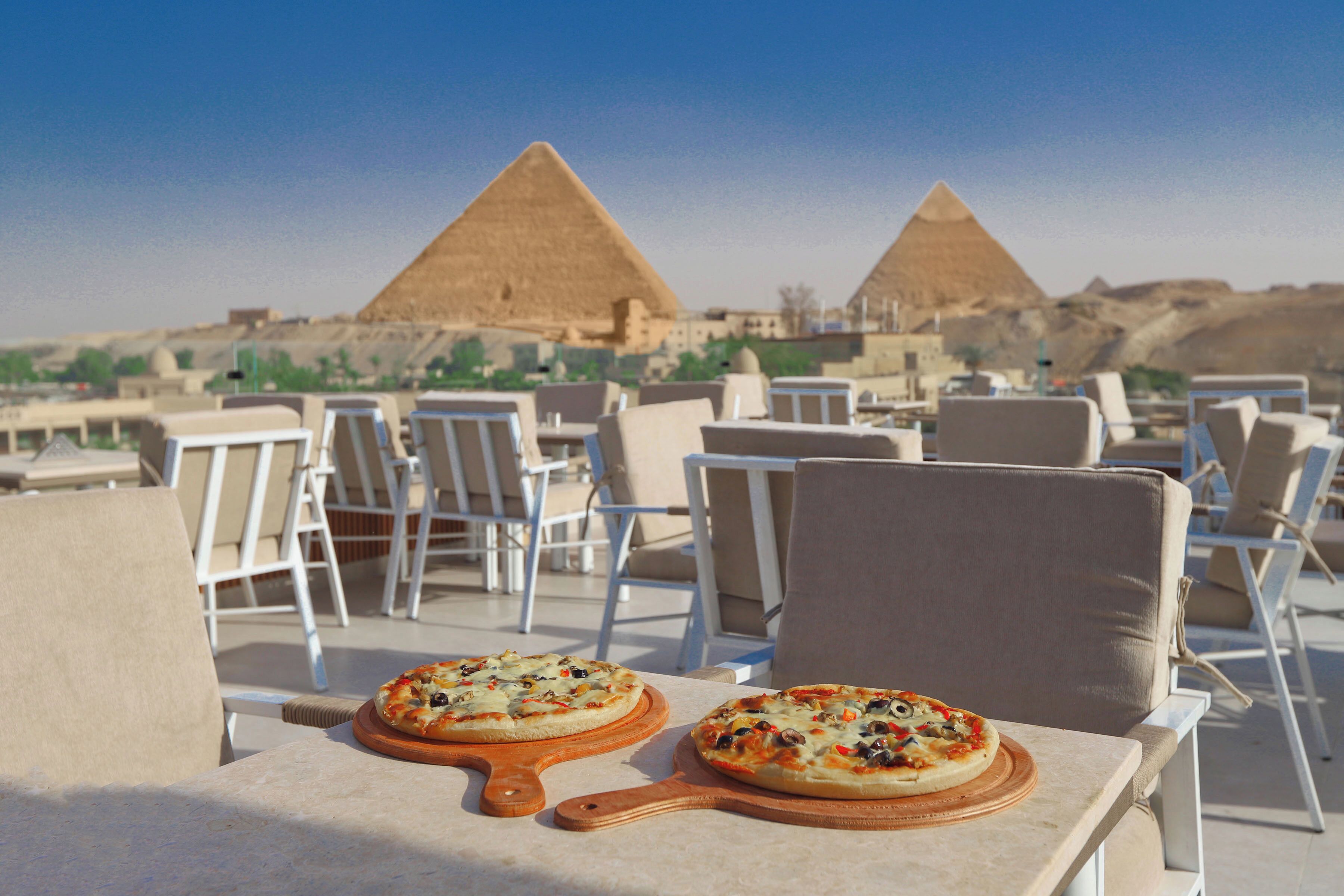 Two pizzas on wooden boards on a table with chairs in the foreground, and pyramids in the background.