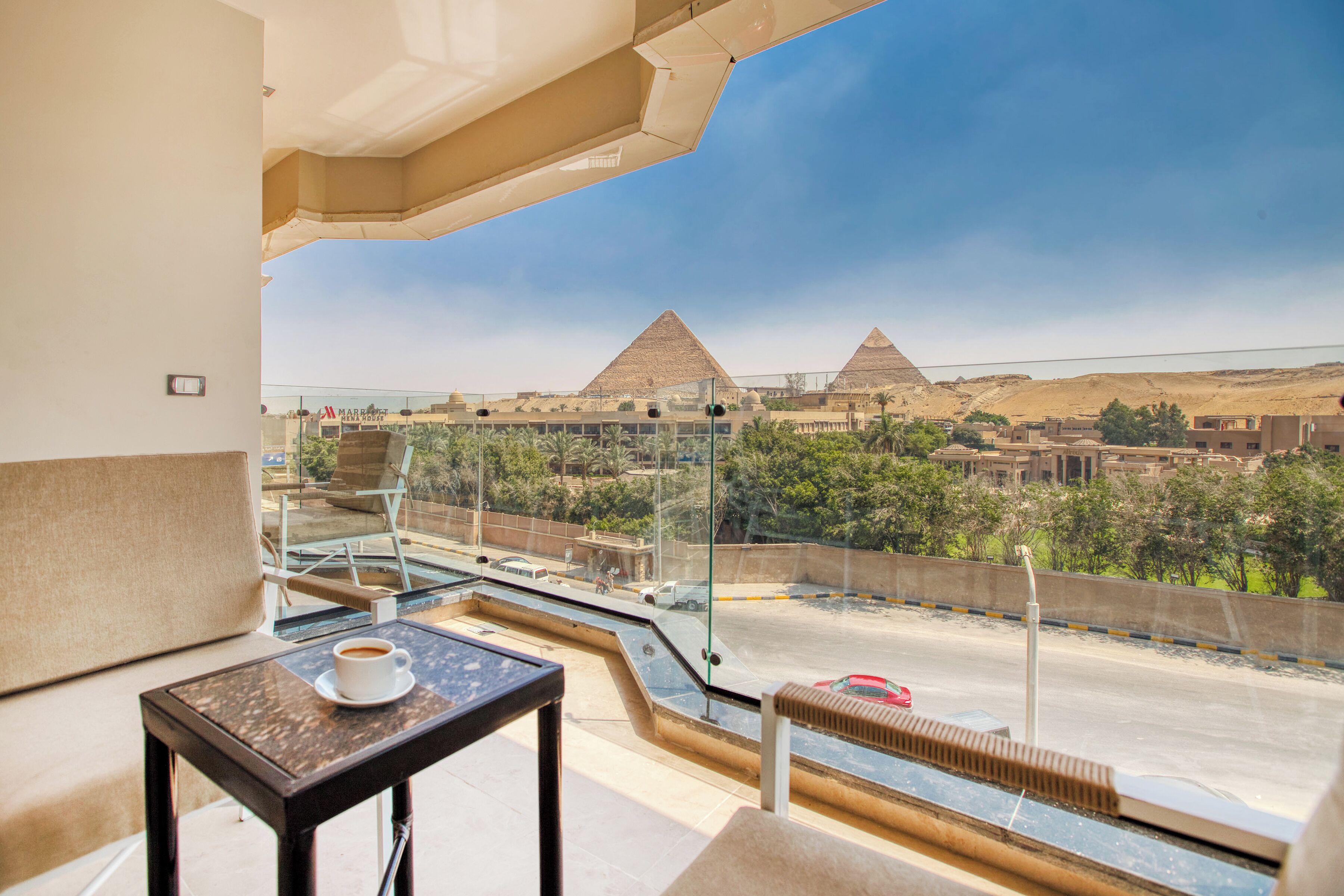 A balcony with a glass railing overlooking a parking area and pyramids in the distance.