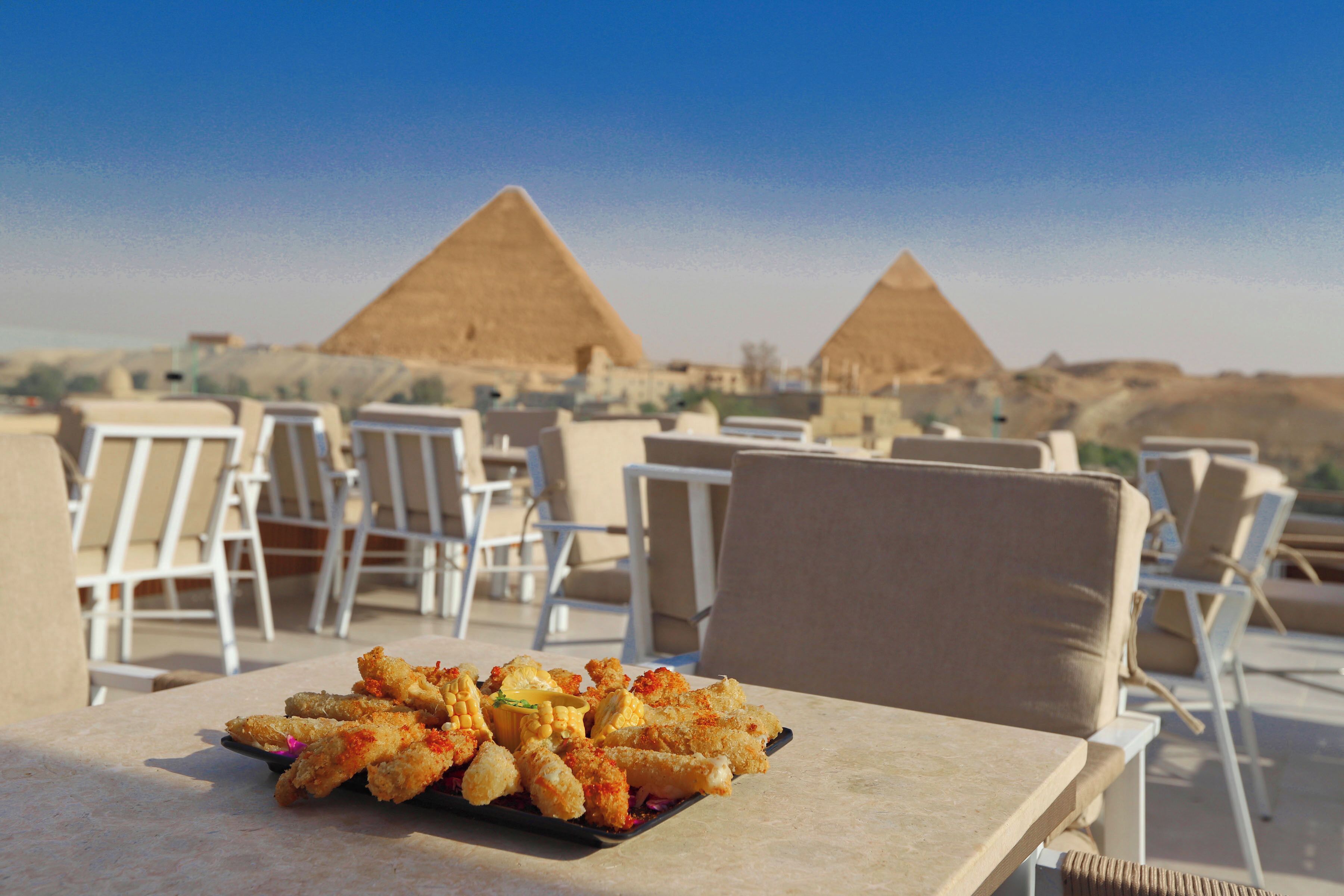 A dining table with a plate of fried food, white lounge chairs, and a view of pyramids in the background.