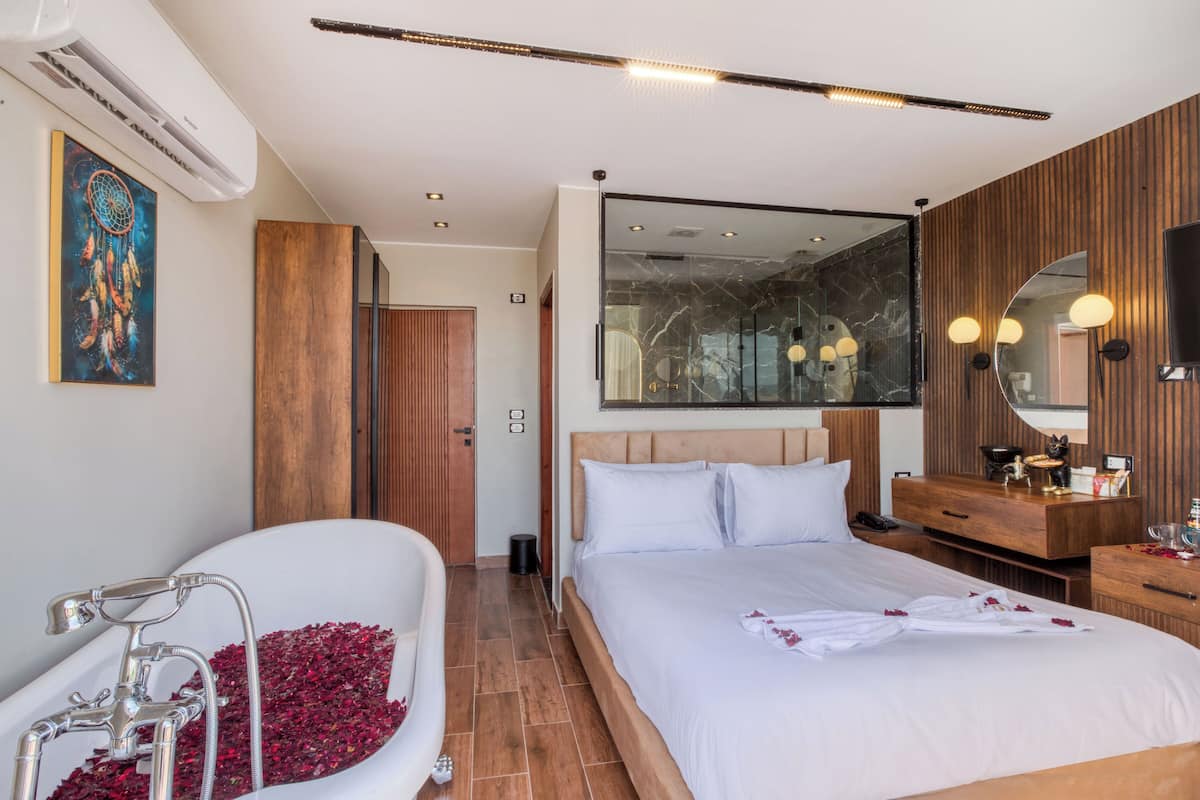 A modern hotel room with a large bed, a separate bathroom with a bathtub, wooden paneling, and a large mirror.