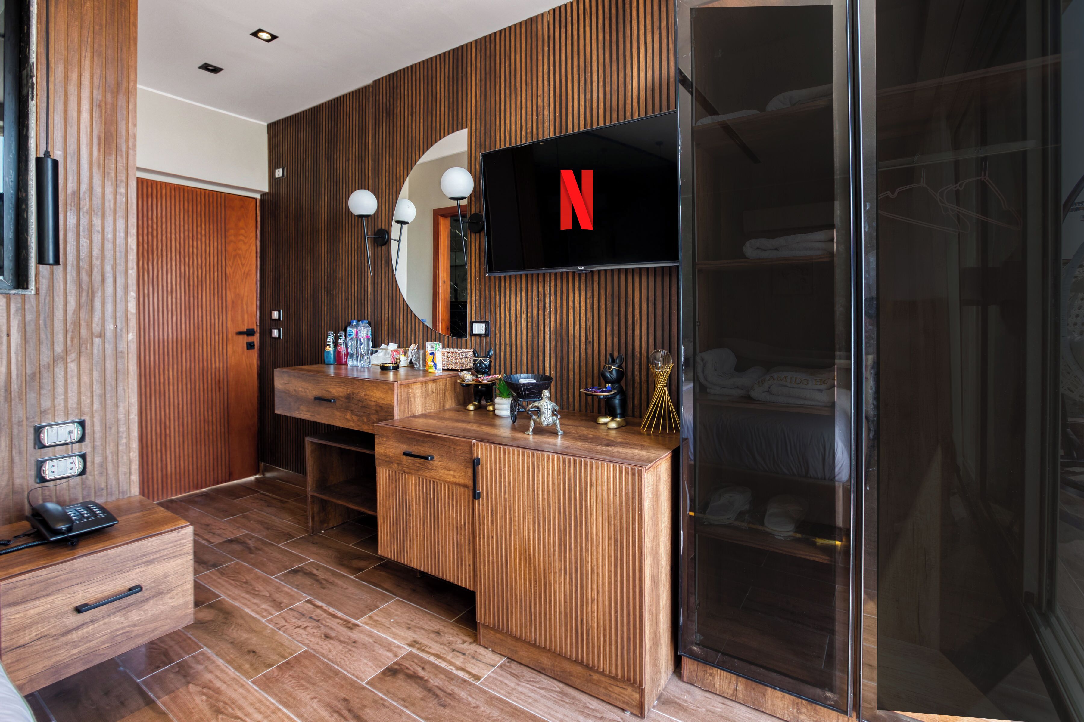 A modern bathroom with wooden paneling, a large mirror, a built-in TV displaying the Netflix logo, and a glass shower enclosure.