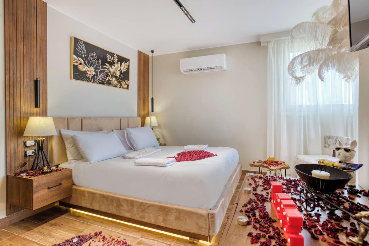 A hotel room with a bed, bedside tables, a wall-mounted air conditioner, a decorative wall piece, and a window with sheer curtains.