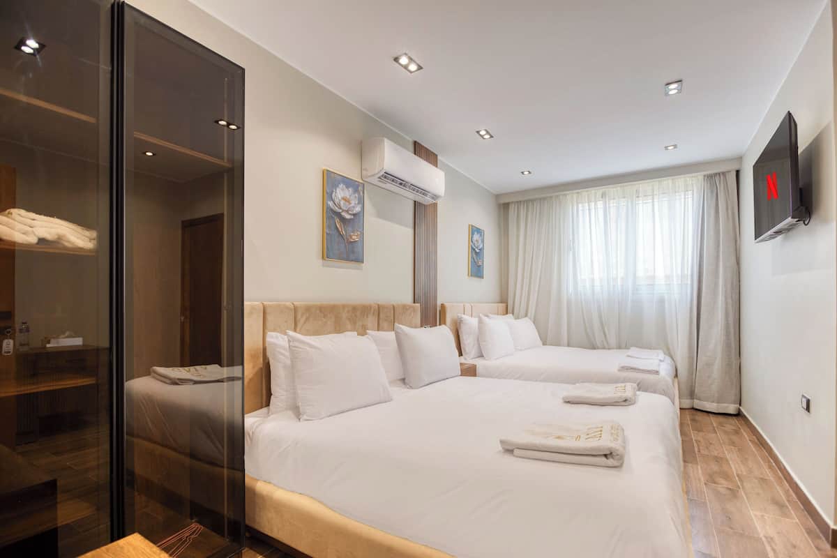 A hotel room with two beds, a TV, an air conditioning unit, and a large window with curtains.