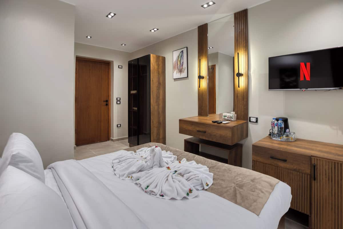 A hotel room with a bed, a wooden wardrobe, a vanity with a mirror, a television mounted on the wall, and a door leading to another room.