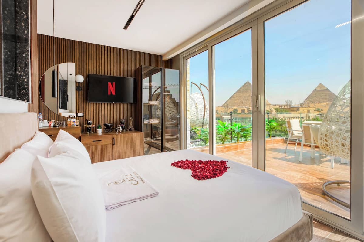 A hotel room with a large bed, a flat-screen TV, a balcony with a view of pyramids, and a dining area with a table and chairs.