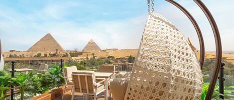 A terrace with a hanging chair, a table, and chairs, overlooking pyramids.