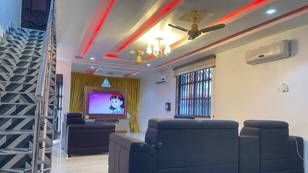 A modern living room with a flat-screen TV, black sofas, a staircase, and a ceiling fan. - Serenity Retreat in Ashongman Estate (North Legon)
