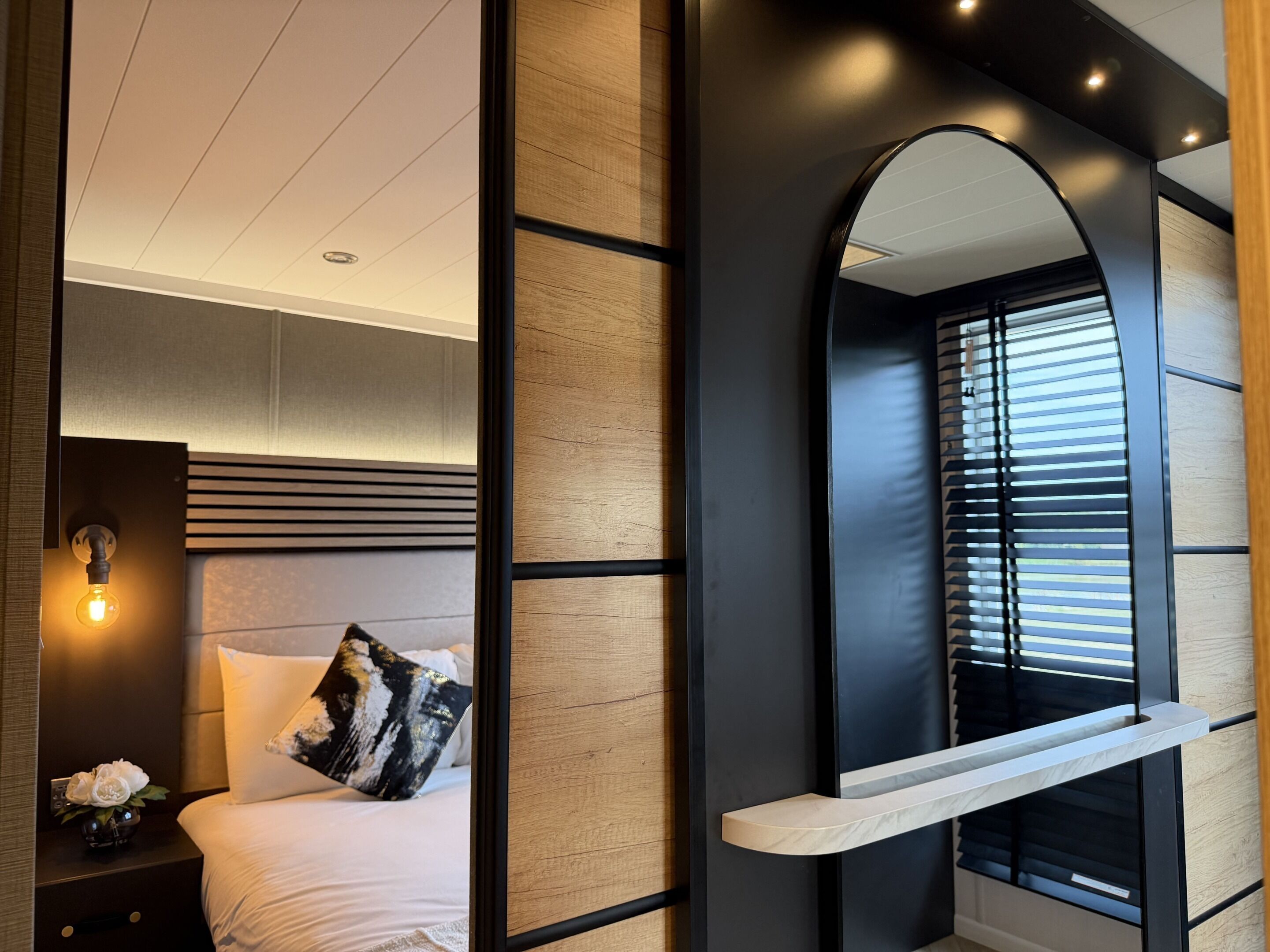 A modern bedroom with a bed, bedside table, lamp, and a large mirror.